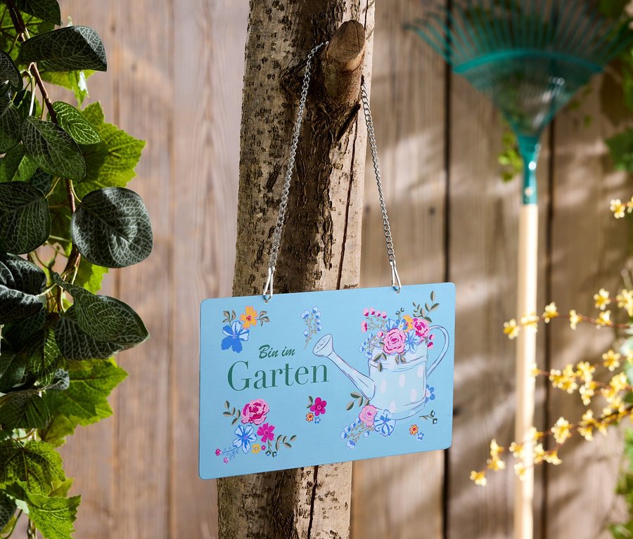 Un panneau bleu avec l'inscription "Bin im Garten" est accroché à un arbre, un râteau en arrière-plan.