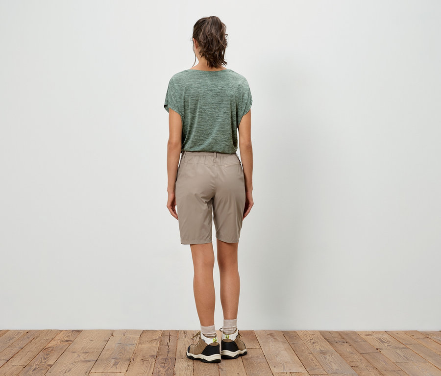 Une femme se tient dos au spectateur, portant un t-shirt vert et un short beige.