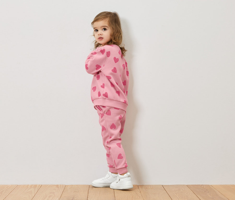 Une petite fille se tient de profil, portant un sweat-shirt rose et un pantalon avec un motif de cœurs, ainsi que des chaussures blanches.