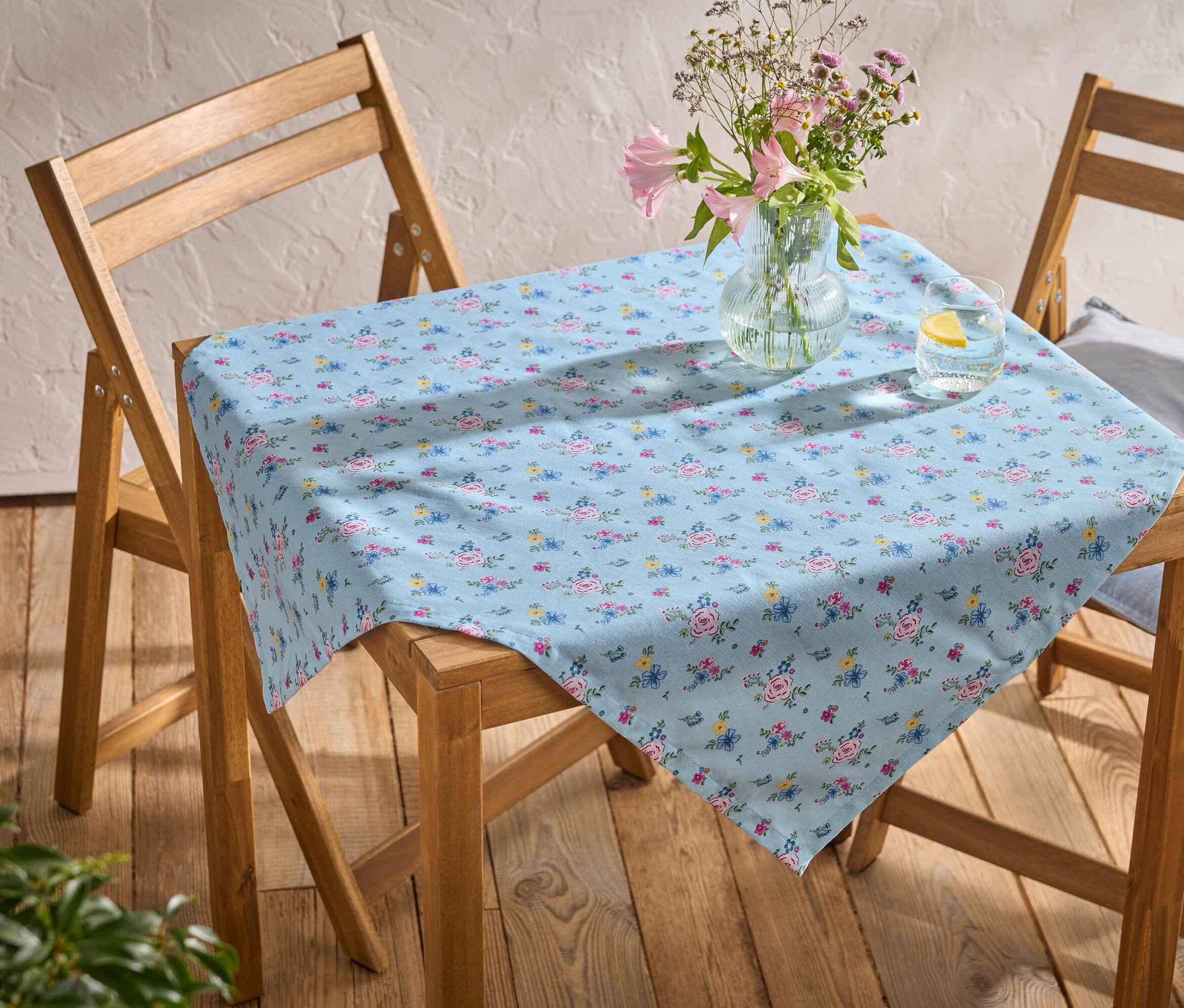 Table avec une nappe bleu clair à motif floral, vase avec des fleurs et verre d'eau avec citron sur deux chaises en bois.