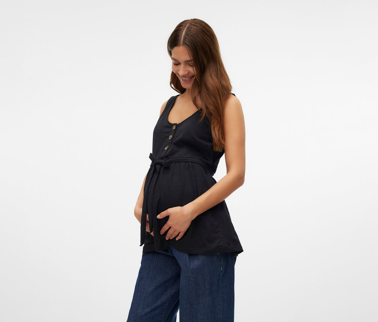 Une femme tient son ventre et porte un top d’allaitement noir MAMALICIOUS «	MLEVI LIA	» et un jean large.
