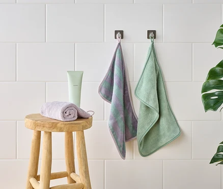 Photo d'un tabouret avec un gant de toilette violet enroulé, un tube vert et deux gants de toilette suspendus devant un mur de carreaux blancs.