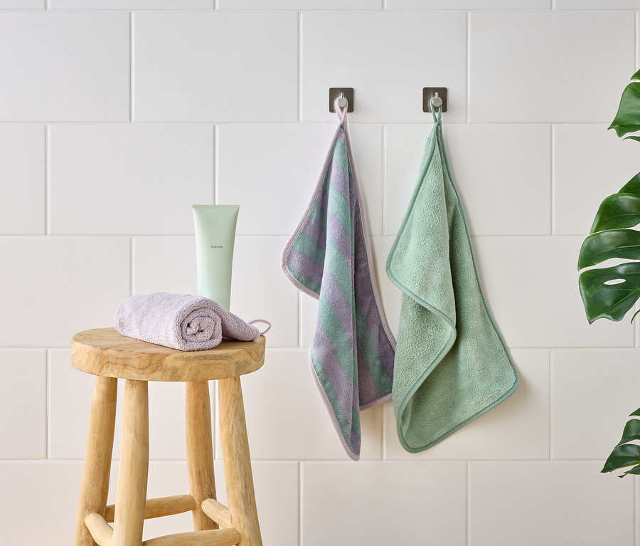 Photo d'un tabouret avec un gant de toilette violet enroulé, un tube vert et deux gants de toilette suspendus devant un mur de carreaux blancs.