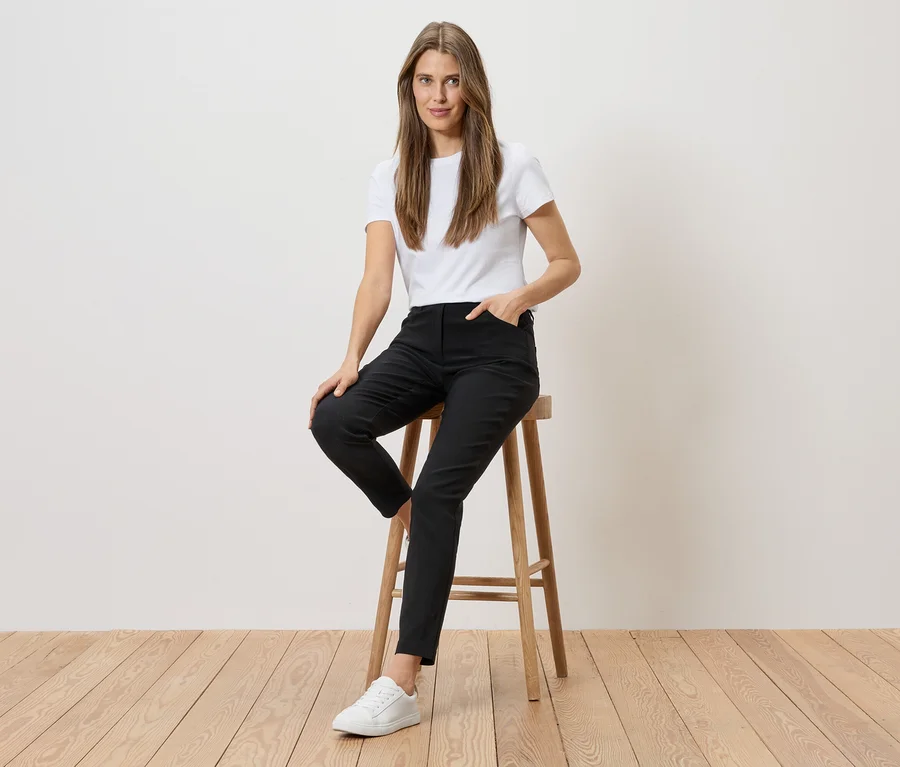 Une femme en t-shirt blanc et pantalon noir est assise sur un tabouret devant un mur blanc et un plancher en bois.