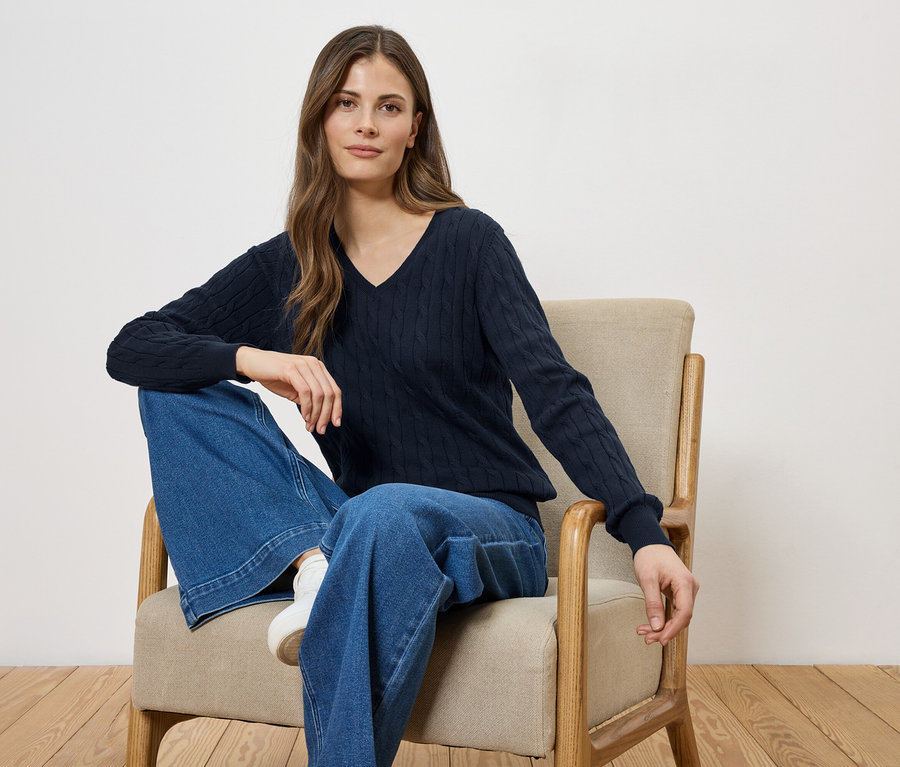 Une femme assise dans un fauteuil, portant un pull bleu marine à col en V et un jean ample.