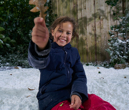 Une jeune fille souriante vêtue d'une veste bleu foncé et d'un pantalon rouge est assise dans la neige, tenant une feuille brune vers le spectateur. Une clôture en bois et de la verdure enneigée sont en arrière-plan.