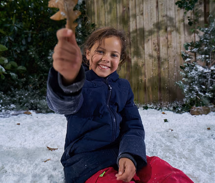 Une jeune fille souriante vêtue d'une veste bleu foncé et d'un pantalon rouge est assise dans la neige, tenant une feuille brune vers le spectateur. Une clôture en bois et de la verdure enneigée sont en arrière-plan.