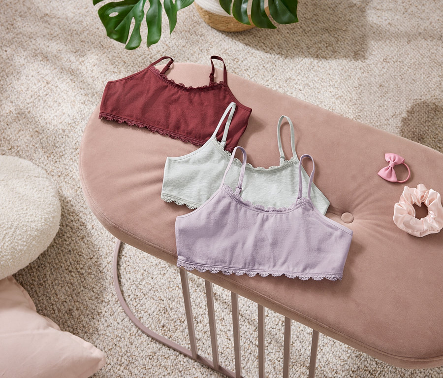Trois bralettes rouge foncé, vert clair et violet clair avec de la dentelle sont posées sur un banc en velours rose. À côté se trouvent un élastique à cheveux rose avec un nœud et un chouchou rose clair. Un coussin blanc et une plante sont visibles en arrière-plan.