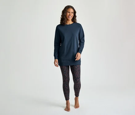 Une femme souriante aux cheveux bruns bouclés se tient de face, vêtue d'un haut tunique bleu marine à manches longues et d'un legging foncé à motifs. Elle est pieds nus.