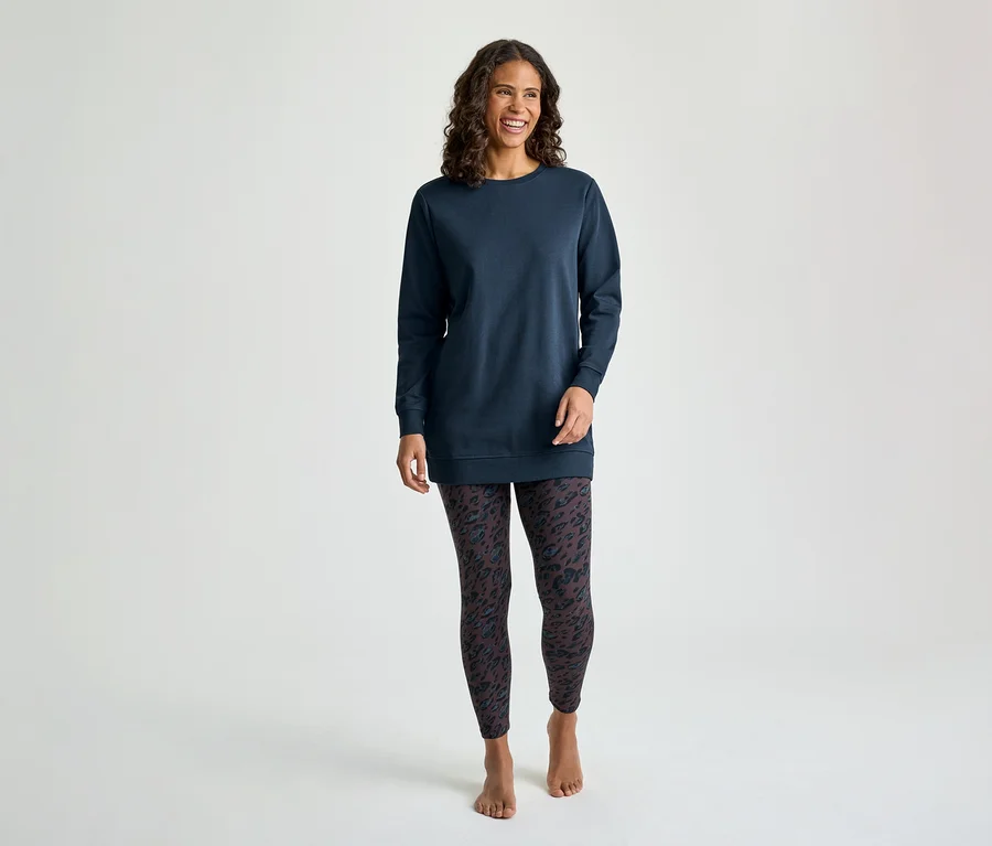 Une femme souriante aux cheveux bruns bouclés se tient de face, vêtue d'un haut tunique bleu marine à manches longues et d'un legging foncé à motifs. Elle est pieds nus.