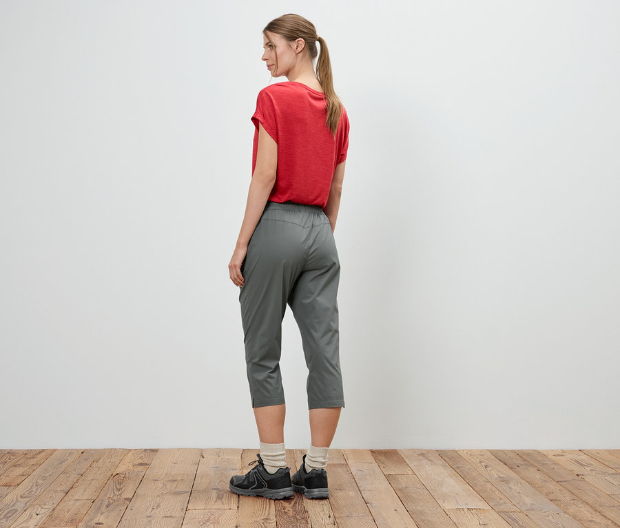 Une femme portant un t-shirt rouge, un pantalon gris et des baskets se tient devant un mur blanc.