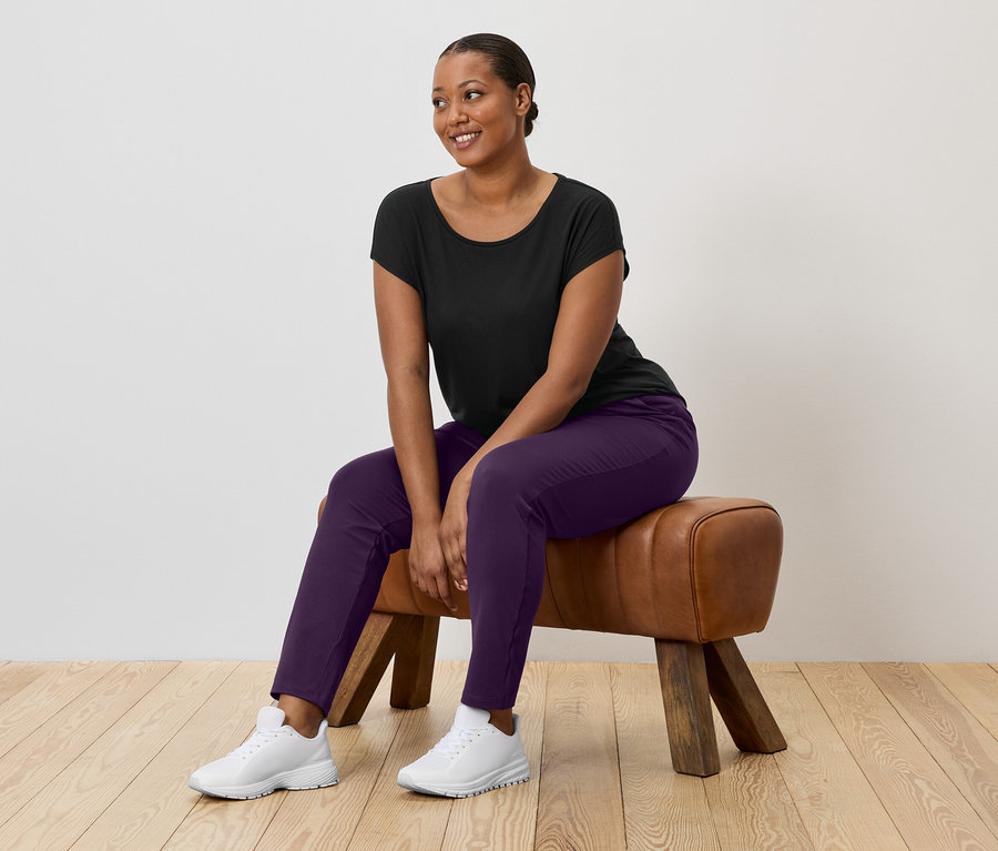 Une femme aux cheveux foncés est assise sur un cheval d'arçons et porte un t-shirt noir, un pantalon violet et des baskets blanches.
