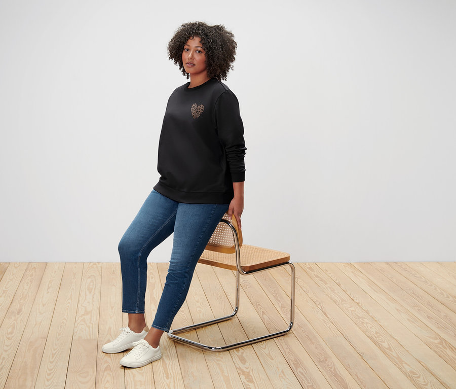 Une femme aux cheveux bouclés est assise sur une chaise, vêtue d'un sweat-shirt noir avec un motif de cœur, d'un jean et de baskets blanches.