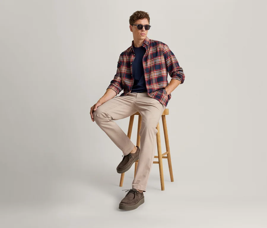 Jeune homme portant des lunettes de soleil, une chemise à carreaux, un t-shirt bleu marine et un pantalon beige, assis sur un tabouret en bois.