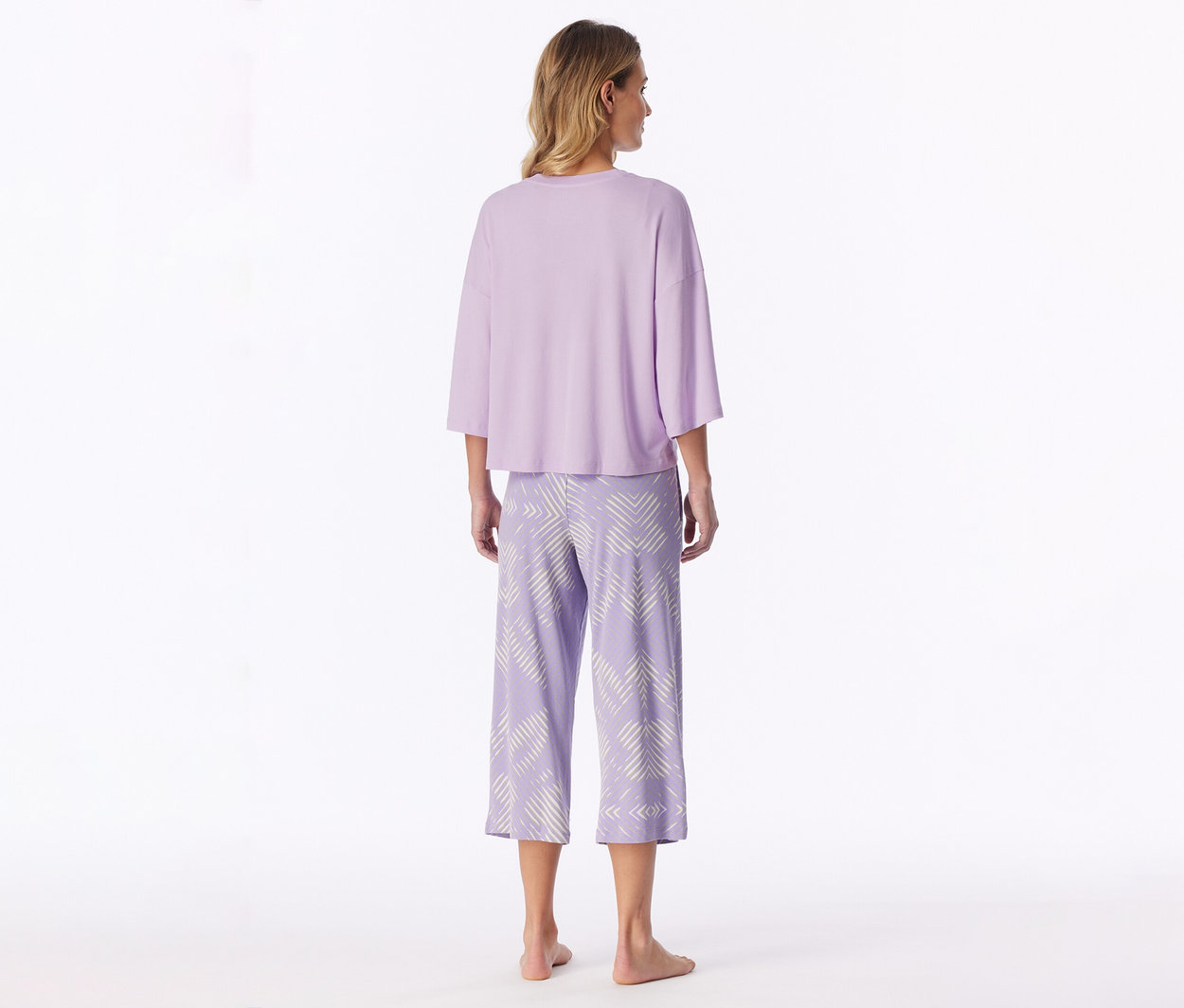 Femme de dos, portant un haut ample violet clair à manches trois-quarts et un pantalon capri violet clair à motifs géométriques, pieds nus sur fond blanc.