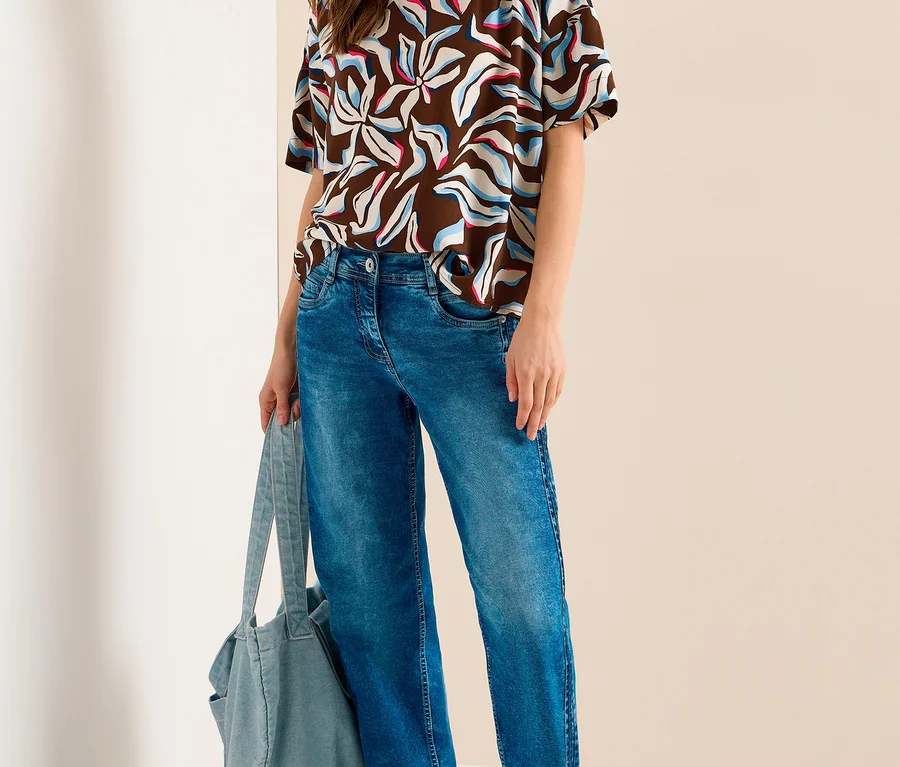 Une femme souriante est debout, vêtue d'un chemisier à manches courtes CECIL marron à motif abstrait blanc, bleu et rose, d'un jean bleu et de baskets argentées, tenant un sac fourre-tout bleu clair.