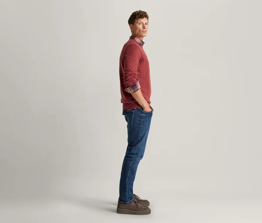 Homme debout de profil, regardant la caméra, portant un pull rouge sur une chemise à carreaux, un jean bleu et des chaussures marron, les mains dans les poches.