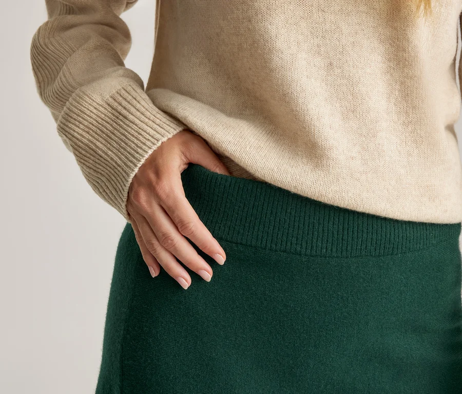 Gros plan d'une personne portant un pull beige à poignets côtelés et une jupe vert foncé à taille côtelée. Une main repose sur la taille de la jupe.