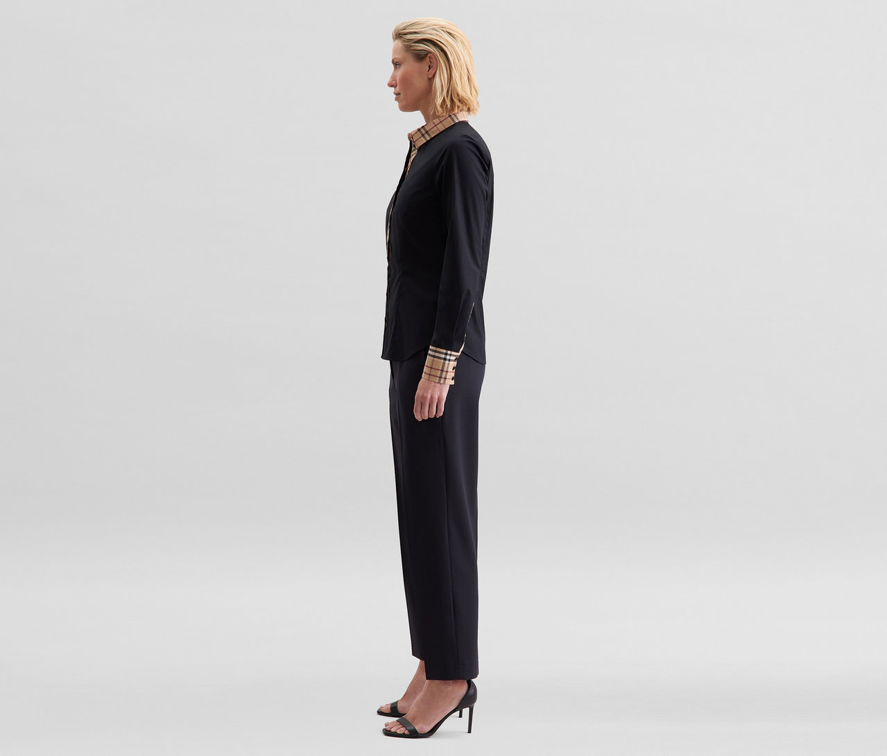 Femme de profil portant un chemisier en popeline à carreaux Seidensticker noir avec col et poignets à carreaux, un pantalon noir et des sandales noires à talons.