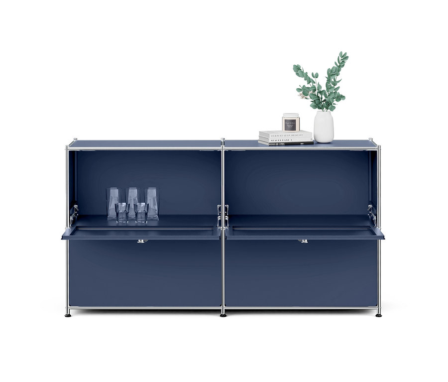 Meuble modulaire bleu foncé avec des cadres chromés et quatre compartiments. Le compartiment supérieur gauche est ouvert, révélant plusieurs verres transparents. Les compartiments inférieurs gauche et droit ont des portes abattantes partiellement ouvertes. Sur le dessus à droite, un vase blanc avec des branches vertes, une pile de trois livres et une petite bougie sont exposés.