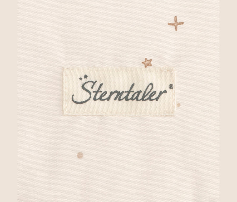 Gros plan d'un tissu beige clair avec une étiquette blanche cousue affichant le logo 'Sterntaler' en gris foncé. De petits motifs marron clair en forme de croix, d'étoile et de point sont dispersés sur le tissu.