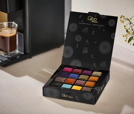 Une machine à café noire Qbo ICONIC Dark Chrome avec un verre de café sur son égouttoir, à côté d'une boîte noire ouverte de Qbo Collection - 16 capsules, révélant 16 dosettes de café colorées. Un vase blanc avec des fleurs blanches et jaunes est en arrière-plan.
