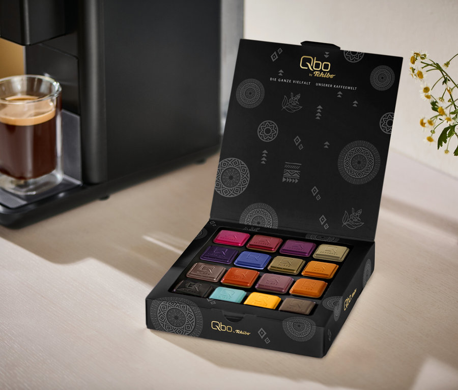 Une machine à café noire Qbo ICONIC Dark Chrome avec un verre de café sur son égouttoir, à côté d'une boîte noire ouverte de Qbo Collection - 16 capsules, révélant 16 dosettes de café colorées. Un vase blanc avec des fleurs blanches et jaunes est en arrière-plan.