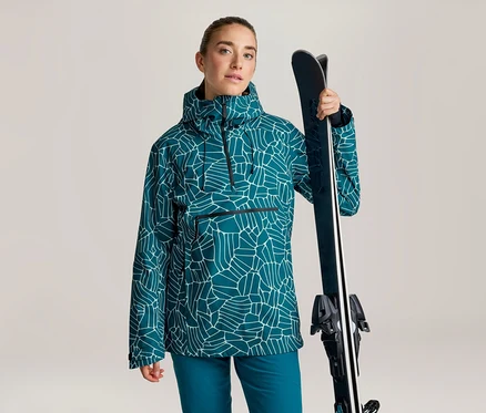Femme portant un anorak turquoise à motif géométrique blanc et un pantalon turquoise, tenant des skis sur son épaule.