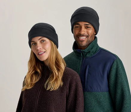 Homme et femme souriants portant des bonnets noirs et des vestes polaires; femme en marron, homme en vert et bleu marine avec poche.