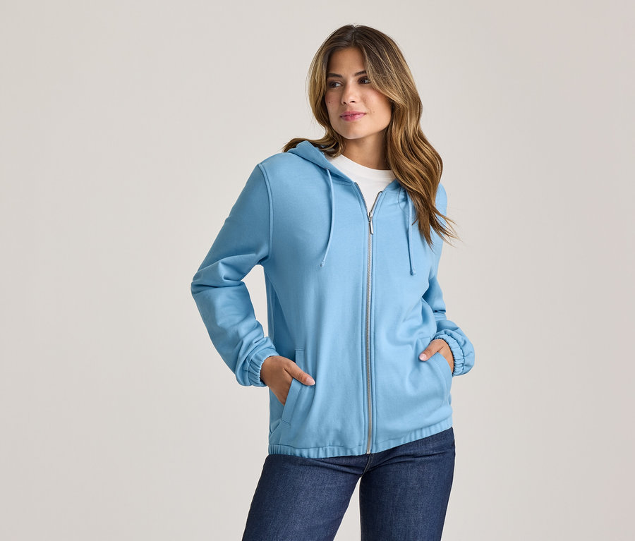 Femme en sweat à capuche zippé bleu clair et jean, les mains dans les poches, regardant vers la droite.