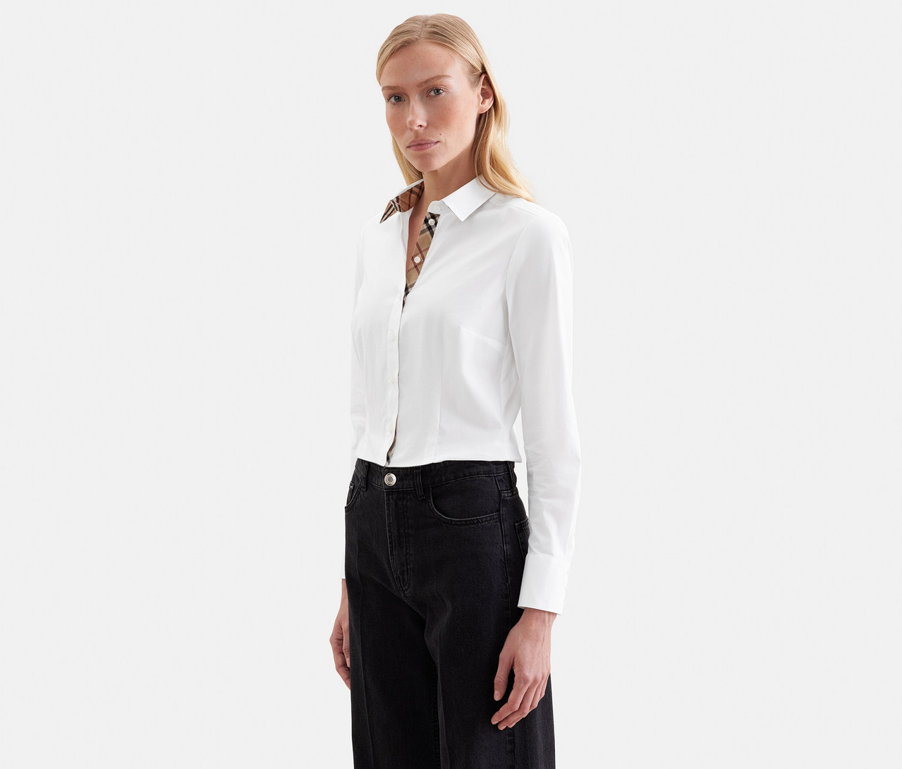 Femme blonde portant une chemise blanche à manches longues avec un col à carreaux et un jean foncé, regardant devant elle.
