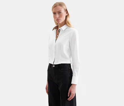Femme blonde portant une chemise blanche à manches longues avec un col à carreaux et un jean foncé, regardant devant elle.