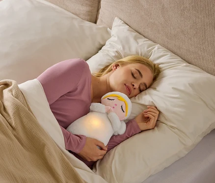 Une femme blonde dort sur le côté dans son lit, vêtue d'un haut rose, serrant une peluche blanche lumineuse au ventre sous une couverture beige.