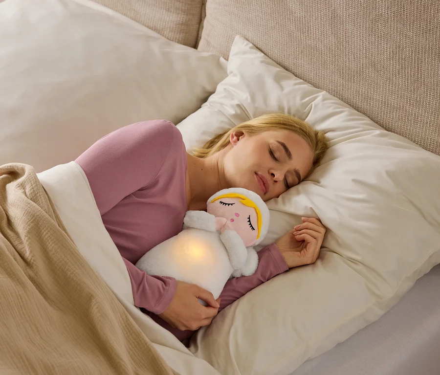 Une femme blonde dort sur le côté dans son lit, vêtue d'un haut rose, serrant une peluche blanche lumineuse au ventre sous une couverture beige.