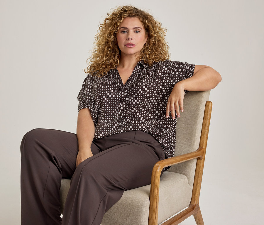 Une femme aux cheveux bouclés est assise sur une chaise claire, vêtue d'un chemisier à manches courtes marron foncé à motifs et d'un pantalon large marron foncé, regardant devant elle.