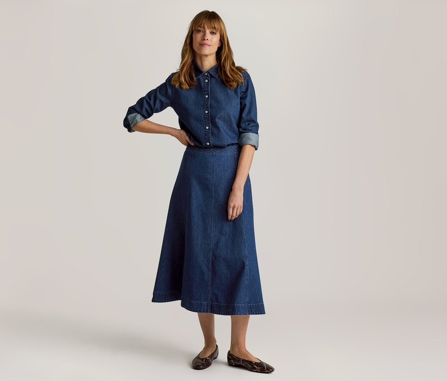 Femme vêtue d'une chemise en jean et d'une jupe midi trapèze, debout, une main sur la hanche, portant des chaussures plates à motifs.