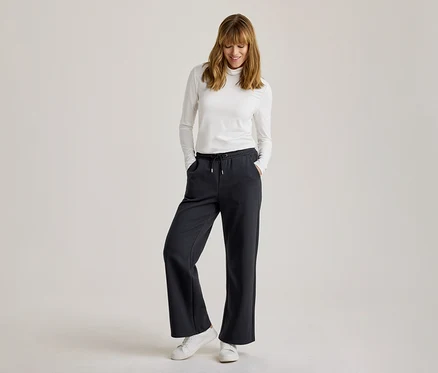 Femme portant un col roulé blanc à manches longues, un pantalon de jogging ample foncé avec cordon de serrage et des baskets blanches, souriant et regardant vers le bas, les mains dans les poches.