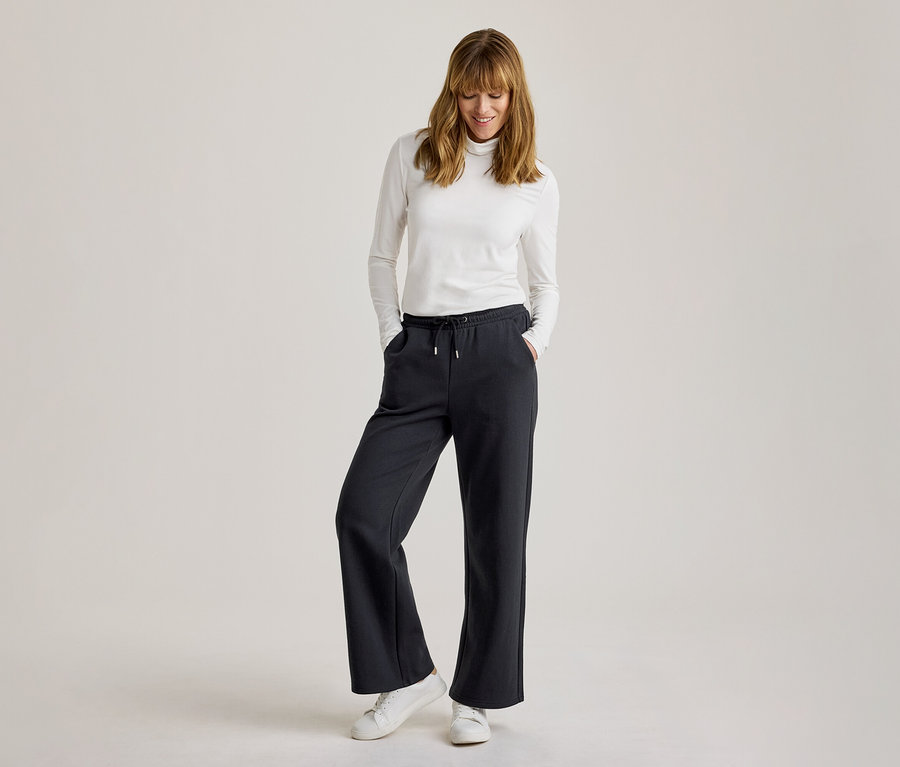 Femme portant un col roulé blanc à manches longues, un pantalon de jogging ample foncé avec cordon de serrage et des baskets blanches, souriant et regardant vers le bas, les mains dans les poches.