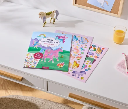 Bureau blanc avec un livre d'autocollants "MEIN STICKERBUCH Einhörner" (plus de 350 autocollants), trois feuilles d'autocollants, une figurine de licorne, un verre de jus d'orange, un carnet rose en peluche, un stylo licorne et un bracelet licorne.