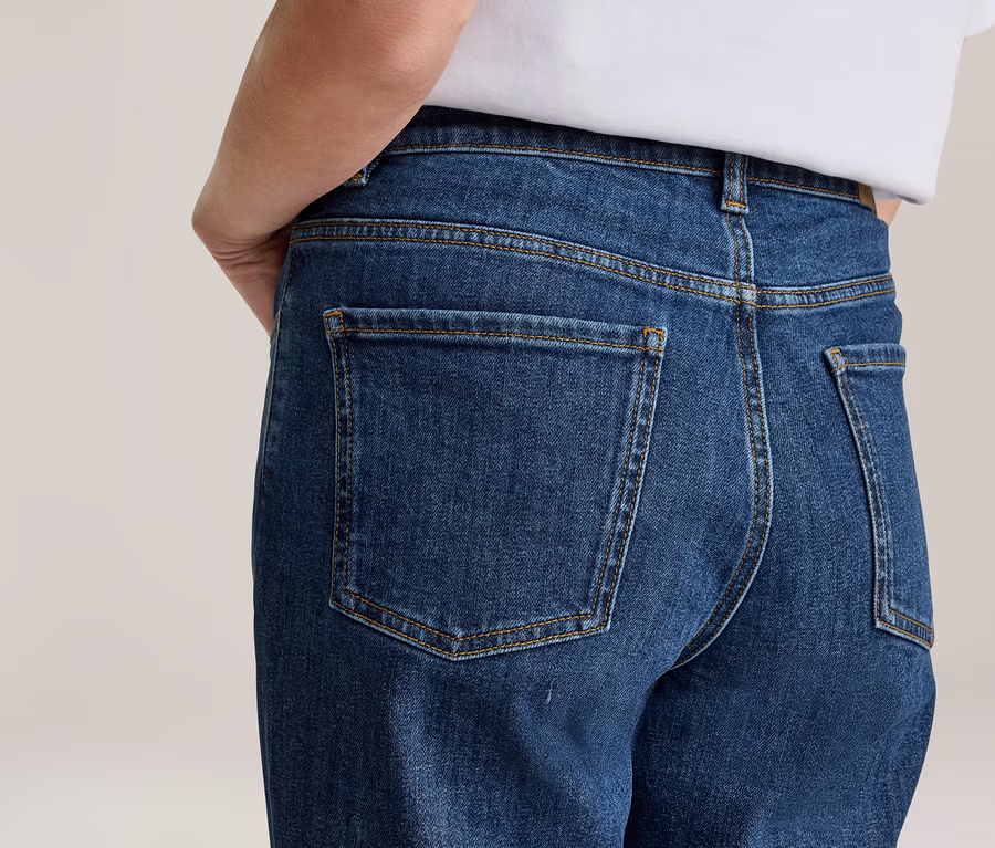 Personne de dos portant un jean bleu et un t-shirt blanc. Le jean a des poches arrière visibles et des surpiqûres jaunes.