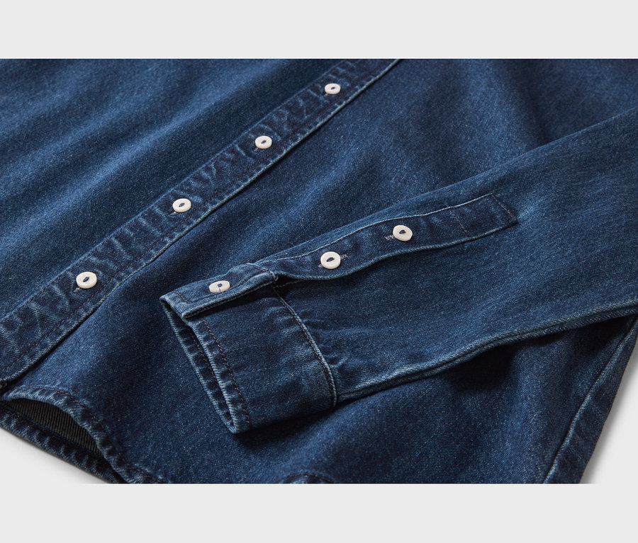 Gros plan d'une chemise en jean bleu foncé, montrant la patte de boutonnage avec des boutons blancs et un poignet de manche également avec des boutons blancs.