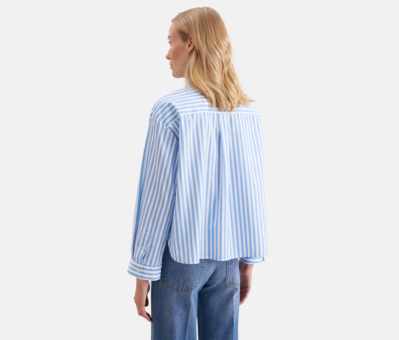 Femme blonde vue de dos, portant un chemisier oversize Seidensticker bleu et blanc à rayures avec des manches longues et un jean bleu.