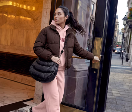 Une femme aux cheveux foncés en queue de cheval ouvre une porte sombre, regardant vers la rue. Elle porte une doudoune marron, un sweat à capuche rose avec le logo FILA, un pantalon de survêtement rose et un sac bandoulière matelassé noir.