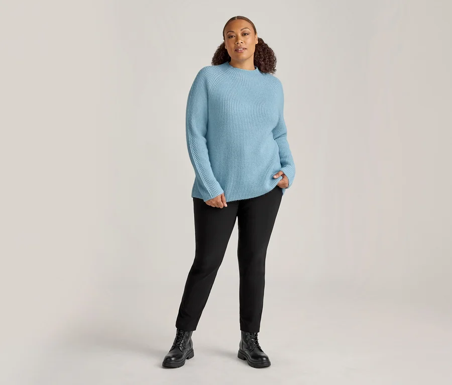 Femme debout, portant un pull côtelé bleu clair et un pantalon en bengaline noir. Sa main gauche est dans sa poche, elle porte des bottes noires.