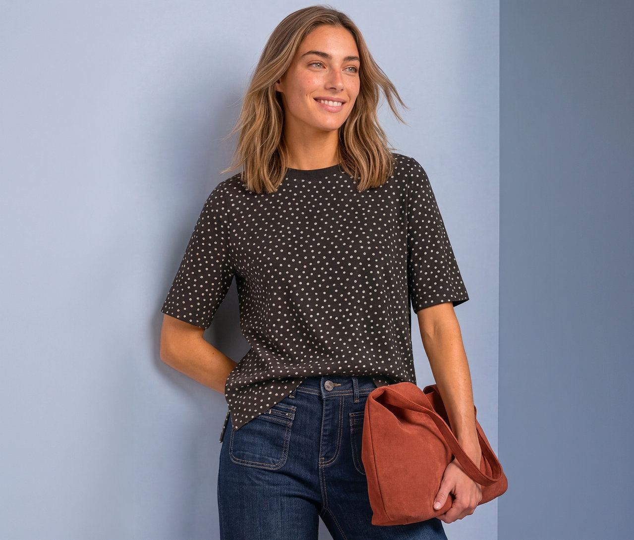 Femme souriante portant un t-shirt foncé à pois blancs et un jean bleu foncé, tenant un sac couleur rouille, devant un fond bleu clair.