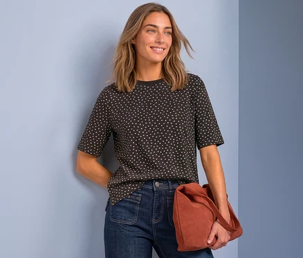 Femme souriante portant un t-shirt foncé à pois blancs et un jean bleu foncé, tenant un sac couleur rouille, devant un fond bleu clair.