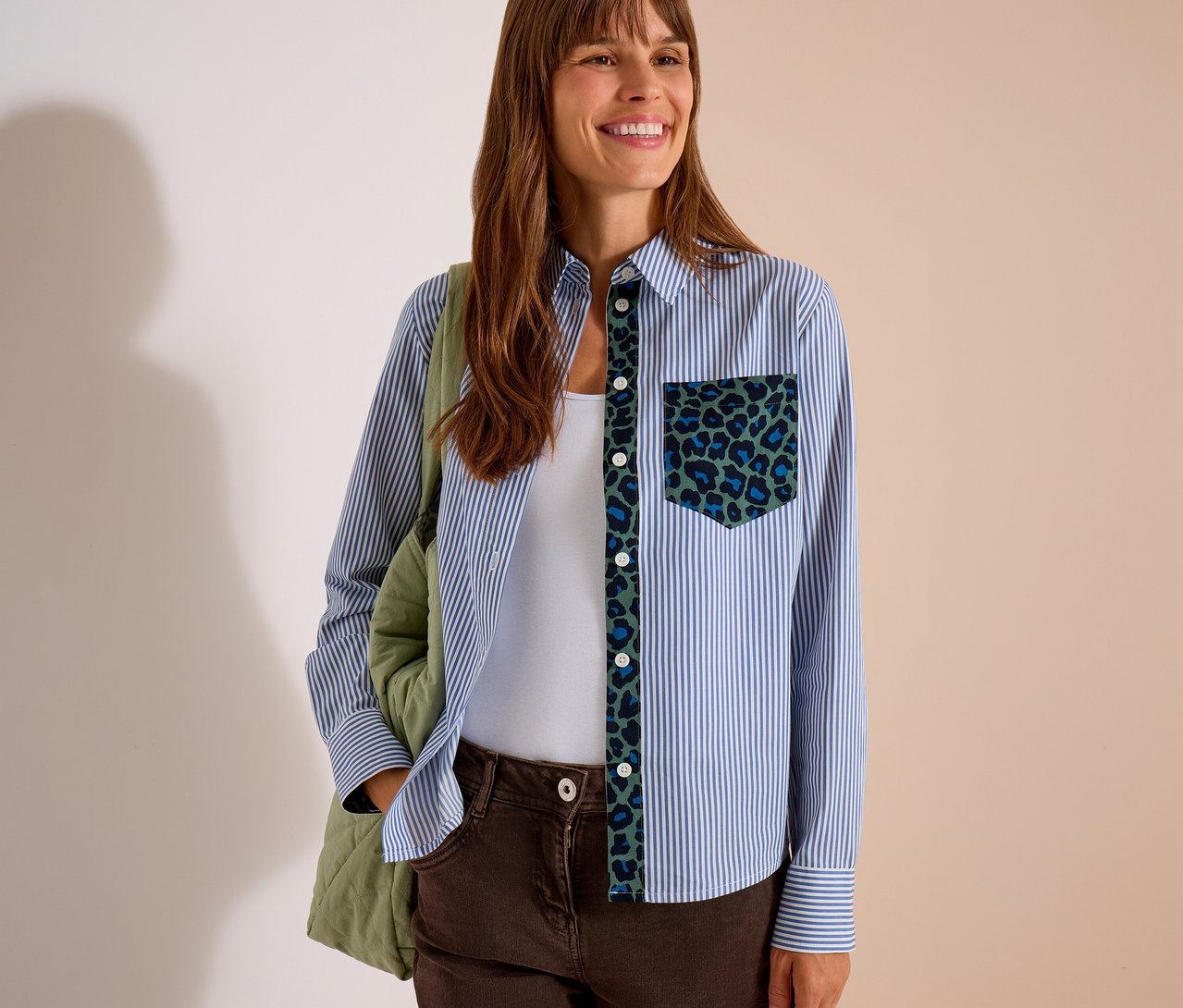 Femme souriante portant un chemisier CECIL rayé bleu et blanc avec un motif léopard vert et bleu sur la patte de boutonnage et la poche, un débardeur blanc et un pantalon marron, avec un sac matelassé vert.