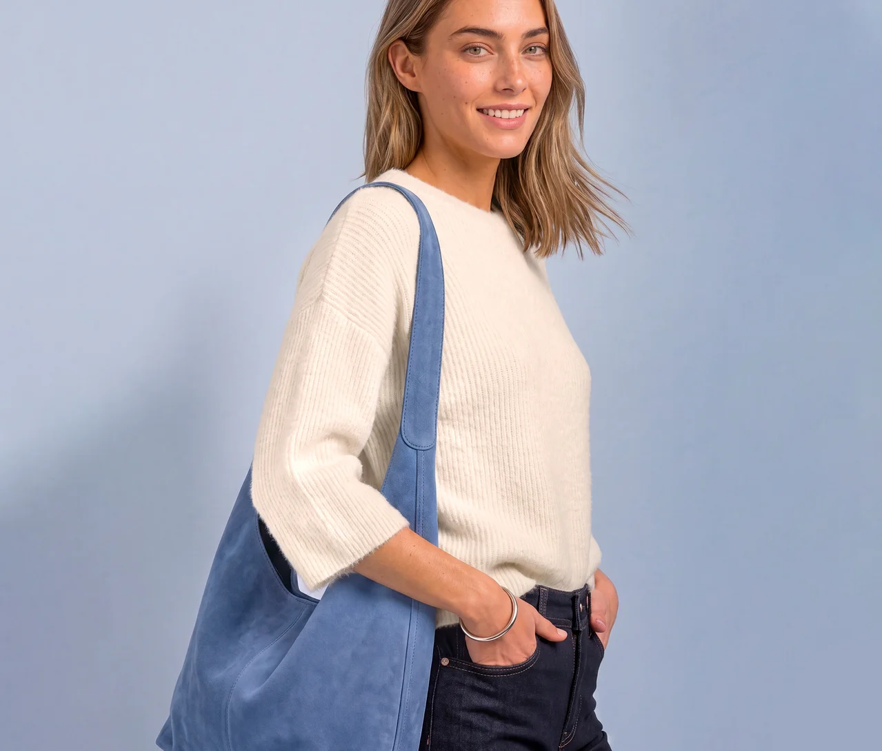 Une femme souriante aux cheveux blonds porte un pull en maille Street One crème, un jean foncé et un sac à bandoulière bleu clair. Sa main droite est dans sa poche.