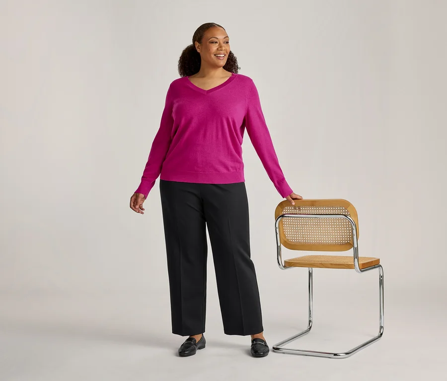 Une femme souriante aux cheveux bouclés foncés, vêtue d'un pull rose à col en V, d'un pantalon noir et de mocassins noirs, se tient à côté d'une chaise en bois clair et chrome, sa main gauche posée sur le dossier.