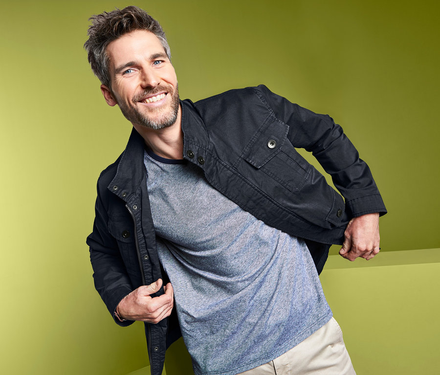 Un homme souriant pose en veste devant un fond vert.